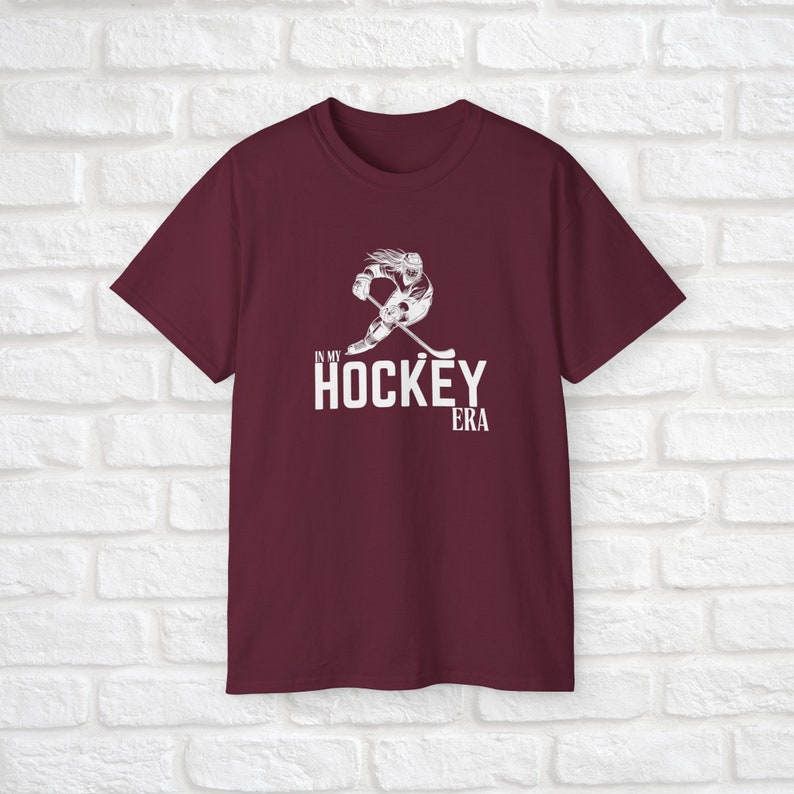 Puede incluir: Camiseta color burdeos con un gr&aacute;fico blanco de un jugador de hockey patinando con un palo y el texto "In My Hockey Era".
