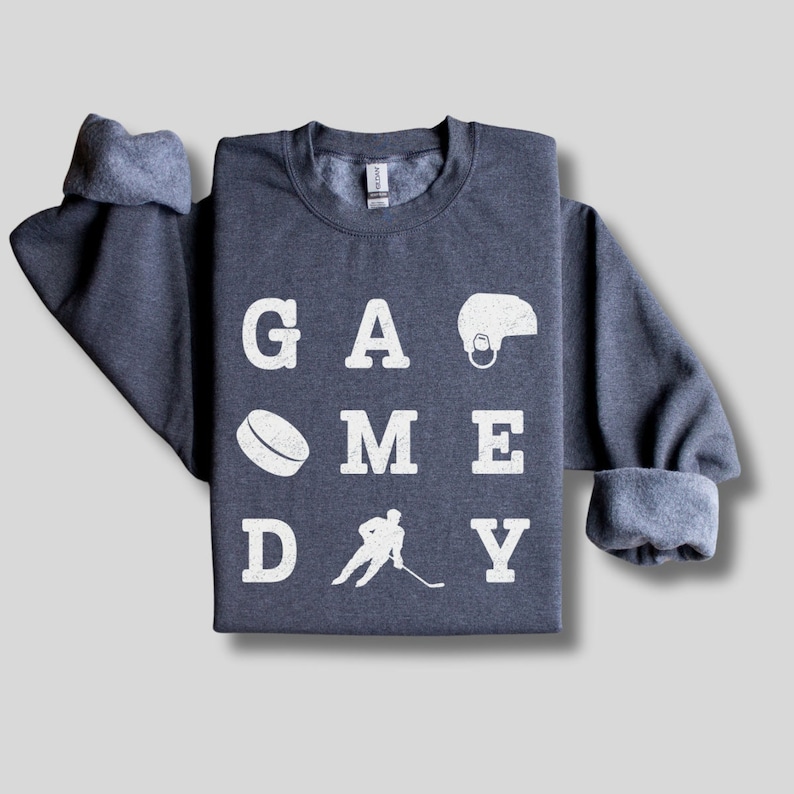 K&ouml;nnte beinhalten: Ein dunkelblauer Pullover mit den Worten "GAME DAY" in wei&szlig;en Buchstaben. Die Buchstaben sind in einem Raster angeordnet, mit einem Hockeypuck, einem Hockeyspieler und einem Hockeyhelm zwischen den Buchstaben.