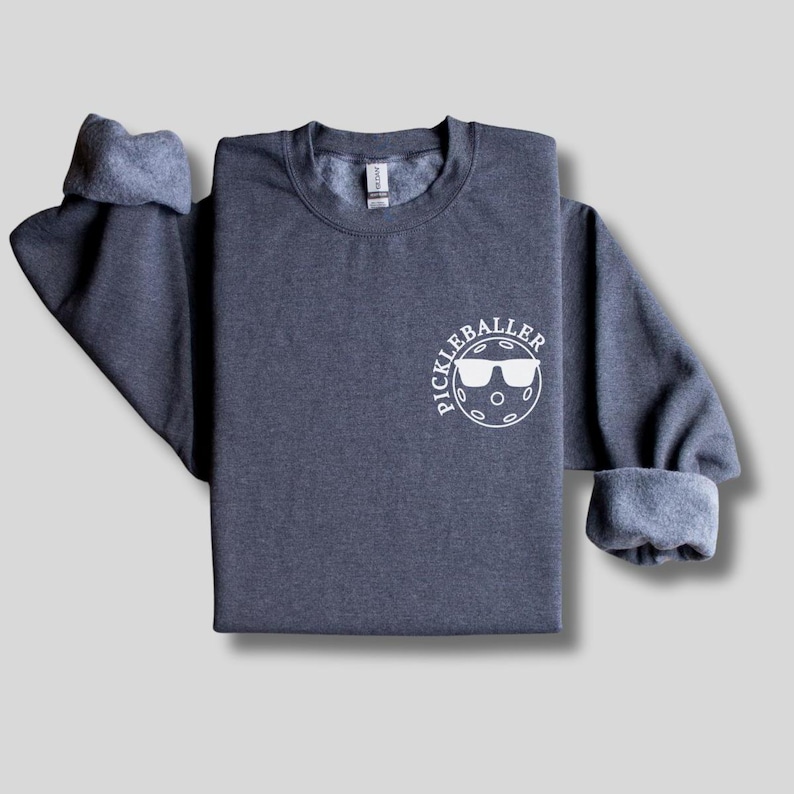 May include: A dark gray sweatshirt with a white circle graphic that says "Pickleballer" with a pickleball and sunglasses inside the circle.