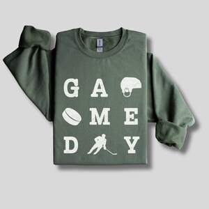 K&ouml;nnte beinhalten: Ein gr&uuml;ner Pullover mit den Worten "GAME DAY" in wei&szlig;en Buchstaben. Die Buchstaben sind in einem Raster angeordnet, mit einem Hockeypuck, einem Hockeyspieler und einem Hockeyhelm zwischen den Buchstaben.