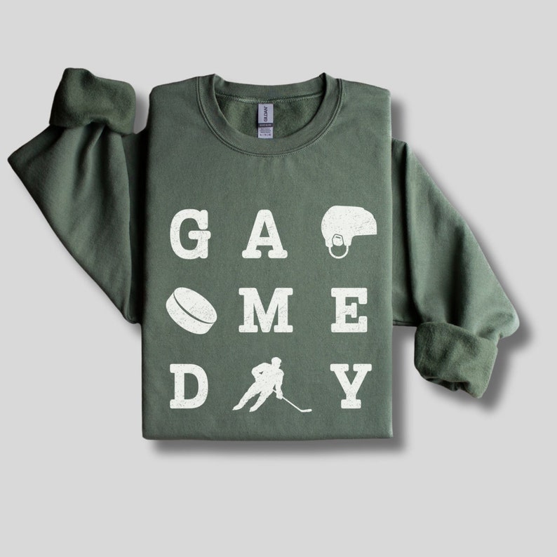 K&ouml;nnte beinhalten: Ein gr&uuml;ner Pullover mit den Worten "GAME DAY" in wei&szlig;en Buchstaben. Die Buchstaben sind in einem Raster angeordnet, mit einem Hockeypuck, einem Hockeyspieler und einem Hockeyhelm zwischen den Buchstaben.