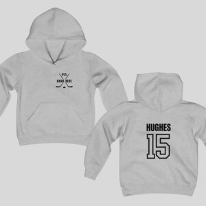 May include: Light gray hoodie with a front pocket and hood. The front features a hockey stick and puck graphic with the text "#15 / NAME HERE". The back displays the name "HUGHES" and the number "15" in black.