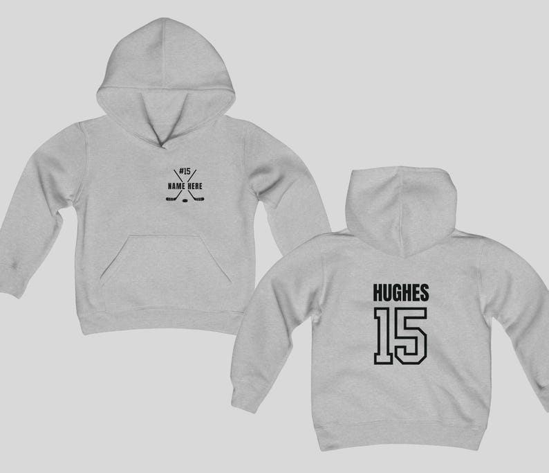 May include: Light gray hoodie with a front pocket and hood. The front features a hockey stick and puck graphic with the text "#15 / NAME HERE". The back displays the name "HUGHES" and the number "15" in black.