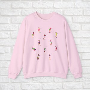 May include: Pink sweatshirt with a graphic print of women playing golf. The print features multiple women in different poses, swinging golf clubs and holding golf balls.