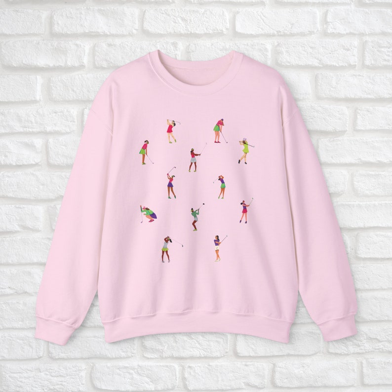 May include: Pink sweatshirt with a graphic print of women playing golf. The print features multiple women in different poses, swinging golf clubs and holding golf balls.