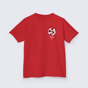 May include: Red short-sleeved t-shirt with a white soccer ball graphic featuring the number 9. The text "Taylor" is written in white below the ball.