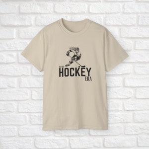 Puede incluir: Una camiseta beige con un gr&aacute;fico negro de un jugador de hockey patinando con un palo. El texto "In My Hockey Era" est&aacute; impreso debajo del gr&aacute;fico.