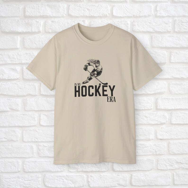 Puede incluir: Una camiseta beige con un gr&aacute;fico negro de un jugador de hockey patinando con un palo. El texto "In My Hockey Era" est&aacute; impreso debajo del gr&aacute;fico.