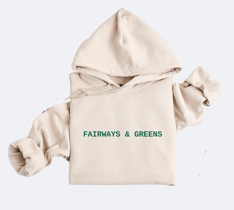 Puede incluir: Una sudadera con capucha beige con un logotipo bordado en verde que dice "FAIRWAYS & GREENS".