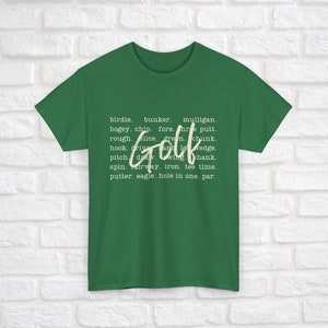 K&ouml;nnte beinhalten: Ein gr&uuml;nes T-Shirt mit dem Wort "Golf" in wei&szlig;er Schrift. Das Wort "Golf" ist umgeben von einer Liste von Golfbegriffen, darunter Birdie, Bunker, Mulligan, Bogey, Chip, Fore, Putt, Rough, Slice, Green, Chunk, Hook, Drive und Low Wedge.