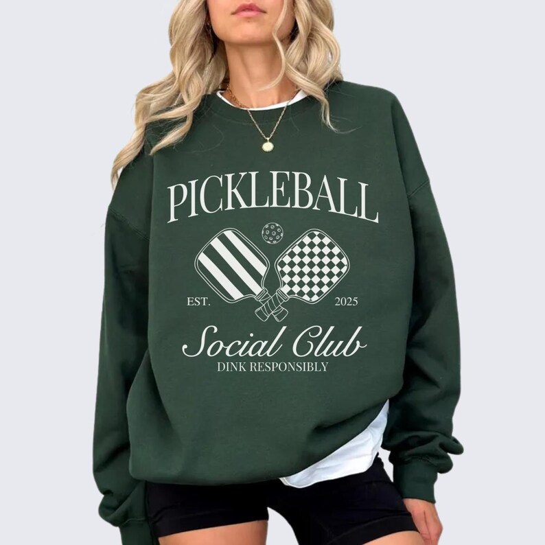 Puede incluir: Sudadera verde con un dise&ntilde;o gr&aacute;fico blanco que dice "Pickleball Social Club Est. 2025 Dink Responsibly" con dos palas de pickleball.