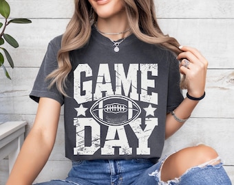 Op de afbeelding: Donkergrijs t-shirt met de woorden "GAME DAY" in grote witte, verweerde letters. Een witte voetbalafbeelding is gecentreerd tussen de woorden, met witte sterren aan weerszijden. Het t-shirt heeft een casual stijl.