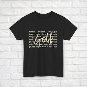 K&ouml;nnte beinhalten: Schwarzes T-Shirt mit dem Wort "Golf" in gro&szlig;en wei&szlig;en Buchstaben. Das Wort ist von einer Liste von Golfbegriffen in kleineren wei&szlig;en Buchstaben umgeben.