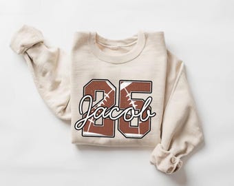 May include: A cream-coloured sweatshirt featuring the number "26" and the name "Jacob" in a football-inspired design. The numbers and name are brown with white detailing, mimicking a football.