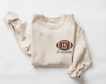 May include: A cream-coloured sweatshirt featuring a football graphic. The graphic shows a brown football with the number 15 and the name "Lucas" in a script font. The sweatshirt is folded.