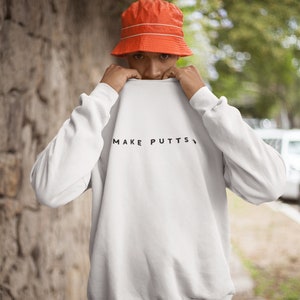 May include: A white sweatshirt with the text "MAKE PUTTS" printed in black on the front. The person is wearing an orange bucket hat.