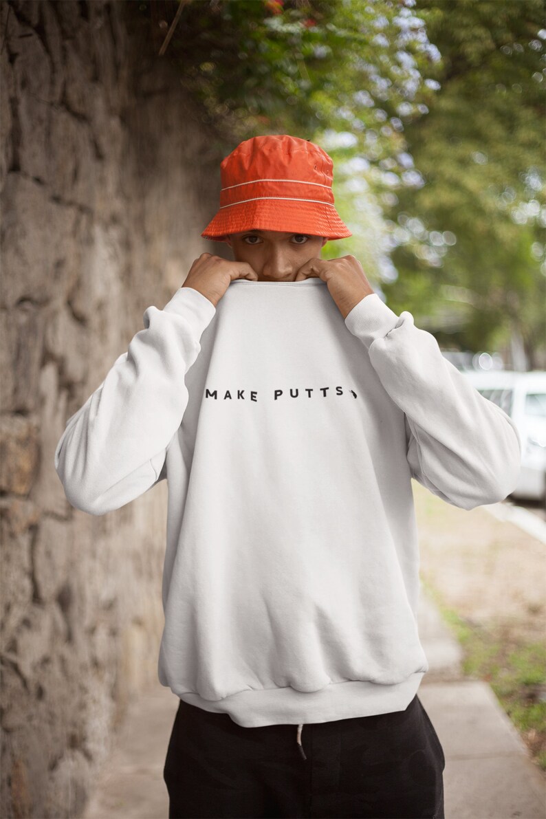 May include: A white sweatshirt with the text "MAKE PUTTS" printed in black on the front. The person is wearing an orange bucket hat.