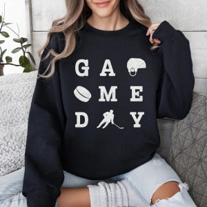K&ouml;nnte beinhalten: Schwarzer Pullover mit wei&szlig;em Text, der "GAME DAY" lautet, mit hockeybezogenen Bildern, darunter ein Hockeyschl&auml;ger, ein Puck und ein Helm.
