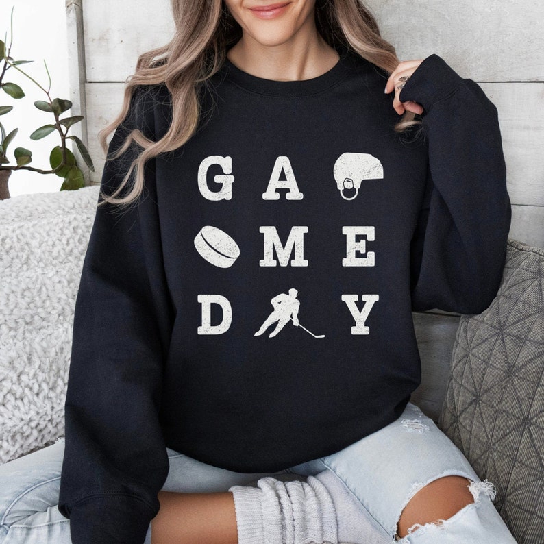 K&ouml;nnte beinhalten: Schwarzer Pullover mit wei&szlig;em Text, der "GAME DAY" lautet, mit hockeybezogenen Bildern, darunter ein Hockeyschl&auml;ger, ein Puck und ein Helm.