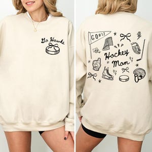 Woman wearing a crewneck sweatshirt with “Hockey Mom” doodle graphic on the back and personalized team name "Go Hawks" and player number "12" on the front; casual hockey game day outfit for moms.