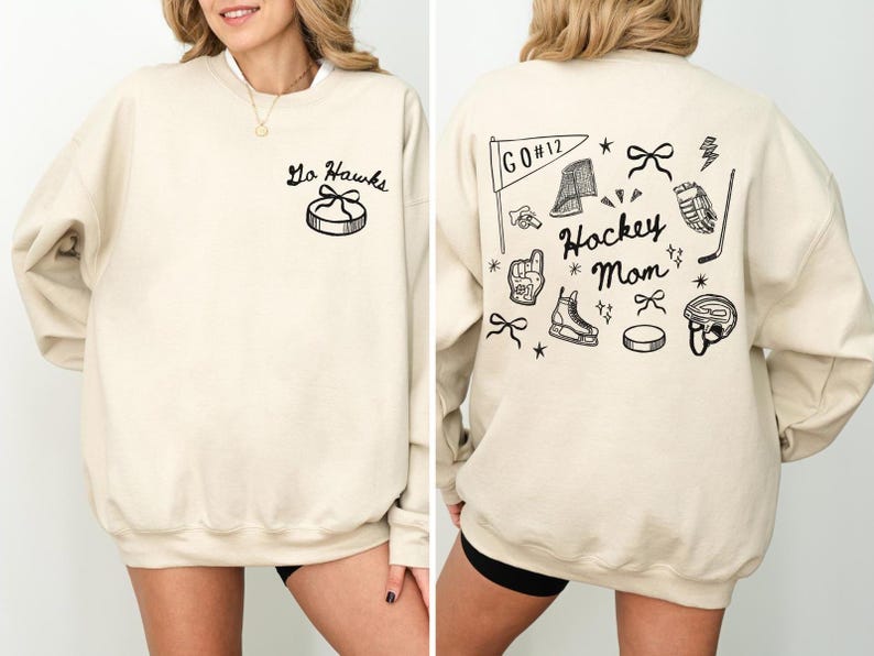 Woman wearing a crewneck sweatshirt with “Hockey Mom” doodle graphic on the back and personalized team name "Go Hawks" and player number "12" on the front; casual hockey game day outfit for moms.