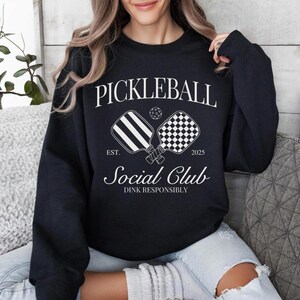 Puede incluir: Sudadera negra con texto blanco que dice "Pickleball Social Club Est. 2023 Dink Responsibly" y un gr&aacute;fico de dos palas de pickleball.