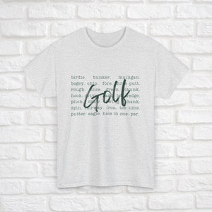 K&ouml;nnte beinhalten: Ein hellgraues T-Shirt mit dem Wort "Golf" in gro&szlig;en gr&uuml;nen Buchstaben. Das Wort "Golf" ist umgeben von einer Liste von Golfbegriffen, darunter Birdie, Bunker, Mulligan, Bogey, Chip, Fore, Three Putt, Rough, Slice, Green, Chunk, Hook, Drive, Wedge, Pitch, Draw, Wing, Shank, Spin, Fairway, Eisen, Tee Time, Putter, Eagle, Hole in One und Par.