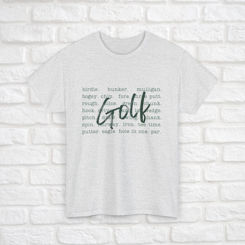 K&ouml;nnte beinhalten: Ein hellgraues T-Shirt mit dem Wort "Golf" in gro&szlig;en gr&uuml;nen Buchstaben. Das Wort "Golf" ist umgeben von einer Liste von Golfbegriffen, darunter Birdie, Bunker, Mulligan, Bogey, Chip, Fore, Three Putt, Rough, Slice, Green, Chunk, Hook, Drive, Wedge, Pitch, Draw, Wing, Shank, Spin, Fairway, Eisen, Tee Time, Putter, Eagle, Hole in One und Par.