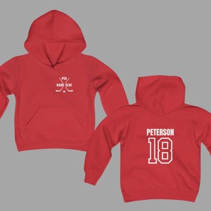 May include: Red hoodie with a front pocket and hood. The front features crossed hockey sticks and the text "#18 NAME HERE". The back displays "PETERSON" and the number "18" in white.