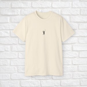 Pu&ograve; includere: Una t-shirt color crema con una piccola silhouette nera di una persona che gioca a pallavolo sul davanti.