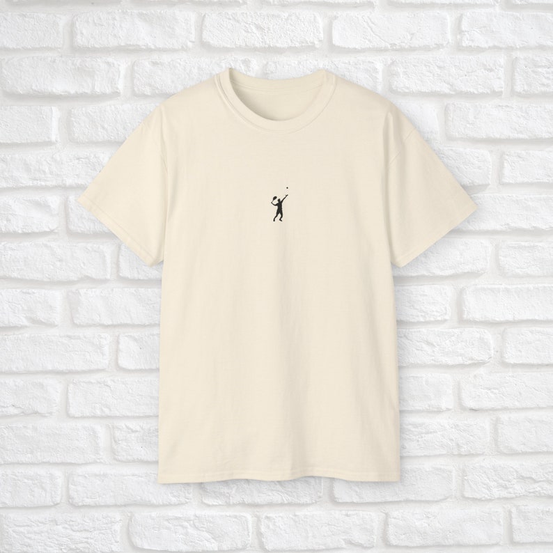 Pu&ograve; includere: Una t-shirt color crema con una piccola silhouette nera di una persona che gioca a pallavolo sul davanti.