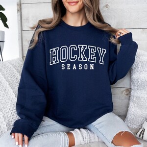 Pu&ograve; includere: Una felpa blu navy con testo bianco che recita "HOCKEY SEASON".