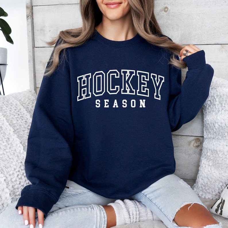 Pu&ograve; includere: Una felpa blu navy con testo bianco che recita "HOCKEY SEASON".