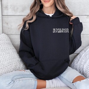 black golf themed hoodie with sunny days and fairways in a bold, trendy font on the back of the shirt. On the front, is a small sunny days and fairways graphic on the left chest