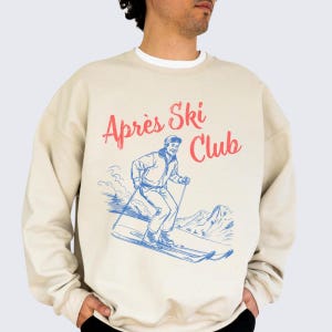 May include: Beige crew neck sweatshirt with a retro graphic. The design shows a skier in blue and white, with the words "Après Ski Club" in red script. The sweatshirt has a vintage ski resort aesthetic.