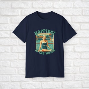 May include: A navy blue t-shirt with a retro-style graphic of a woman in a swimsuit sitting by a pool. The graphic says "Happiest in the Water".