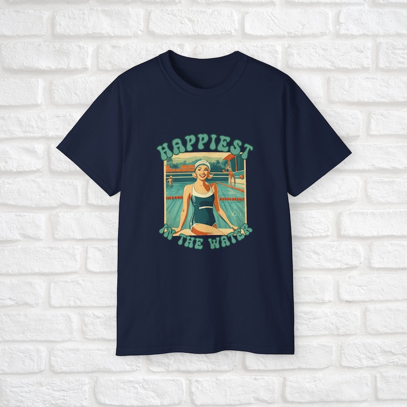 May include: A navy blue t-shirt with a retro-style graphic of a woman in a swimsuit sitting by a pool. The graphic says "Happiest in the Water".
