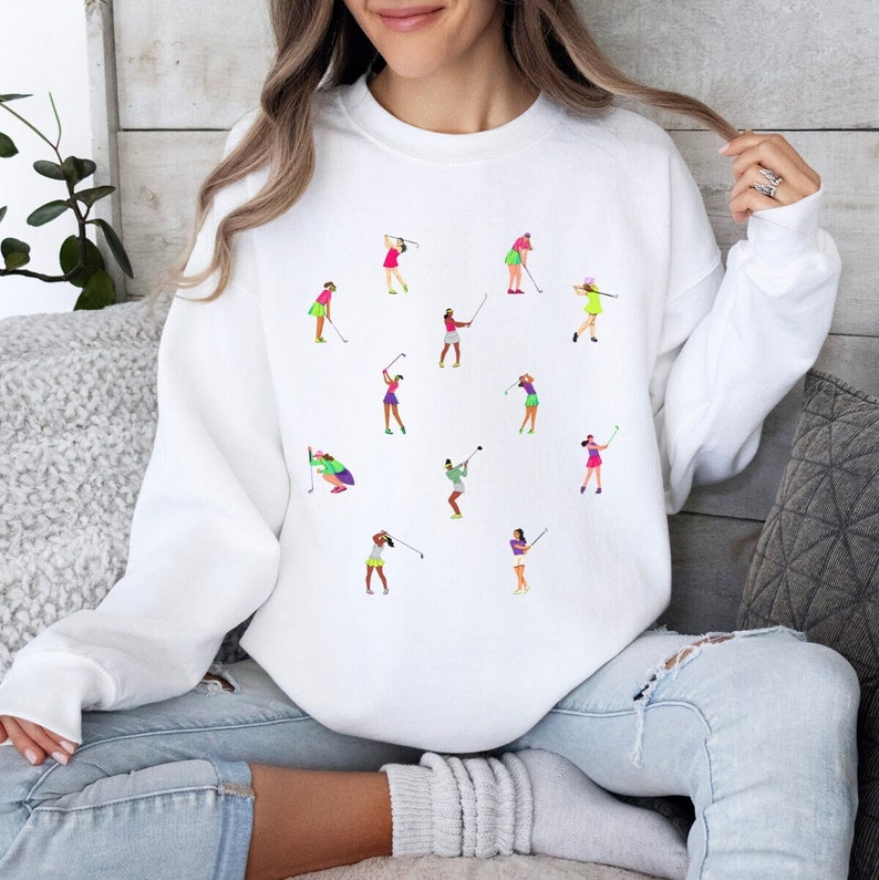 May include: White sweatshirt featuring a colourful graphic print of women playing golf. The women are wearing different coloured clothing and are in various poses.