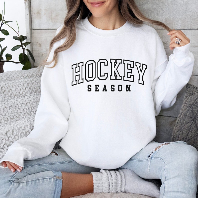 Pu&ograve; includere: Felpa bianca con testo nero che recita "HOCKEY SEASON" in un font in stile universitario.