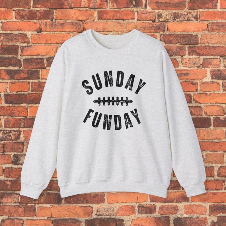 Op de afbeelding: Een lichtgrijs sweatshirt met de tekst "SUNDAY FUNDAY" in zwart, met een voetbalafbeelding tussen de woorden.