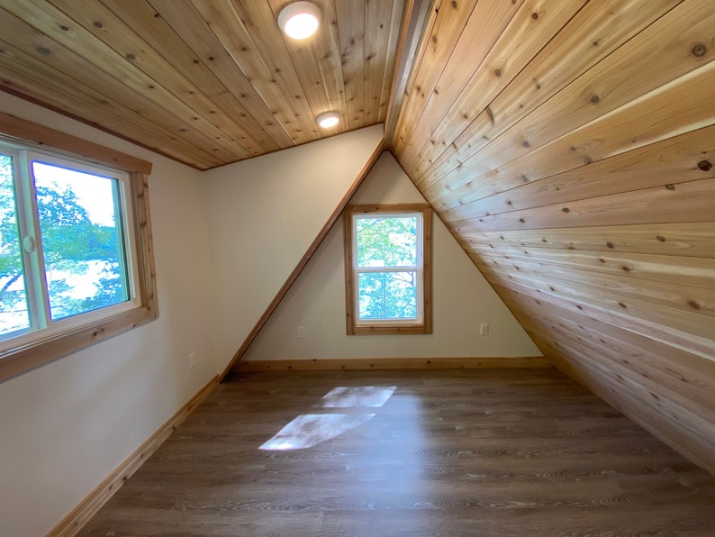 May include: A room with a sloped ceiling and wood paneling. There are two windows, one on each side of the room. The floor is a light brown wood.