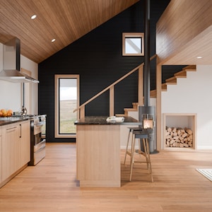 Puede incluir: Una cocina y sala de estar modernas con gabinetes, pisos y techo de madera clara. Una pared de acento negro contrasta con la madera clara. Se ven una estufa y una campana extractora de acero inoxidable. Una escalera conduce a un nivel superior.