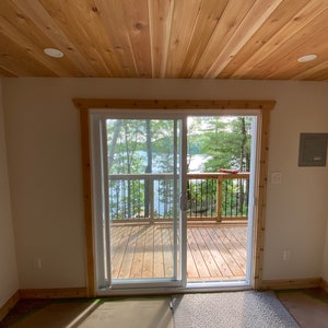 May include: A white sliding glass door with a wooden frame leads to a wooden deck overlooking a lake and trees. The room has a wood paneled ceiling and white walls.
