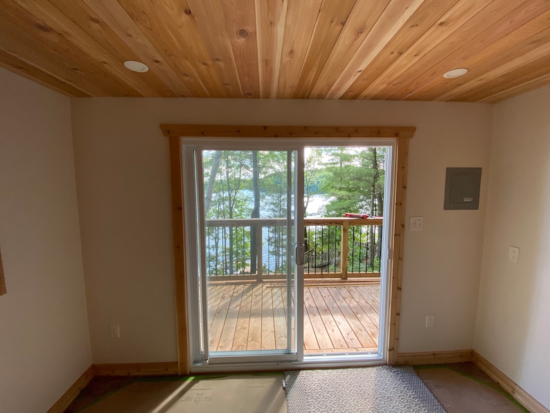 May include: A white sliding glass door with a wooden frame leads to a wooden deck overlooking a lake and trees. The room has a wood paneled ceiling and white walls.