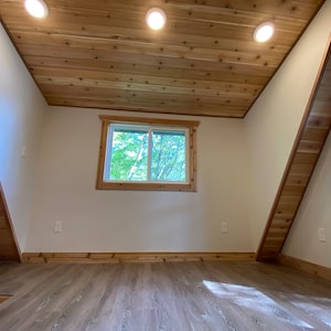 May include: A room with a sloped ceiling and wood paneling. The room has a large window with a view of trees outside. The floor is made of light brown wood.