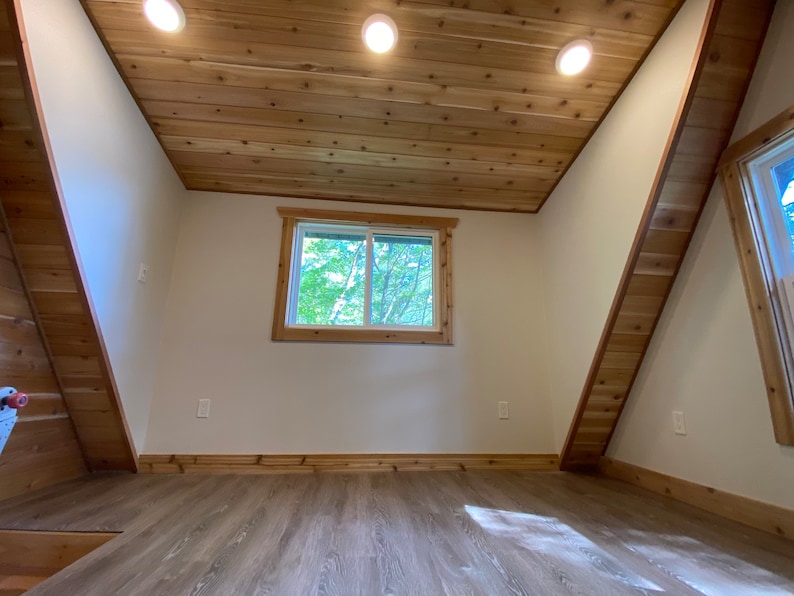 May include: A room with a sloped ceiling and wood paneling. The room has a large window with a view of trees outside. The floor is made of light brown wood.