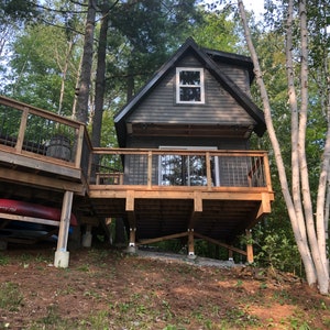 May include: A small, dark gray cabin with a wooden deck and a window. The cabin is built on stilts and is surrounded by trees.