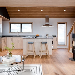 Puede incluir: Interior de cocina moderno con gabinetes de madera clara, un refrigerador de acero inoxidable y una encimera negra. La habitaci&oacute;n cuenta con una isla de cocina con dos taburetes, una campana extractora de acero inoxidable y una estufa negra. Una chimenea negra y una mesa de centro de cristal completan el espacio.