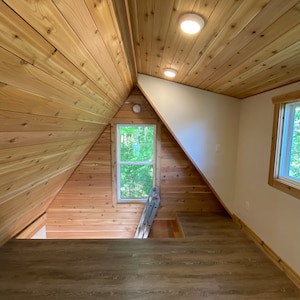 May include: A loft space with wood paneling on the walls and ceiling. There is a window with a view of trees outside. A ladder is leaning against the wall.