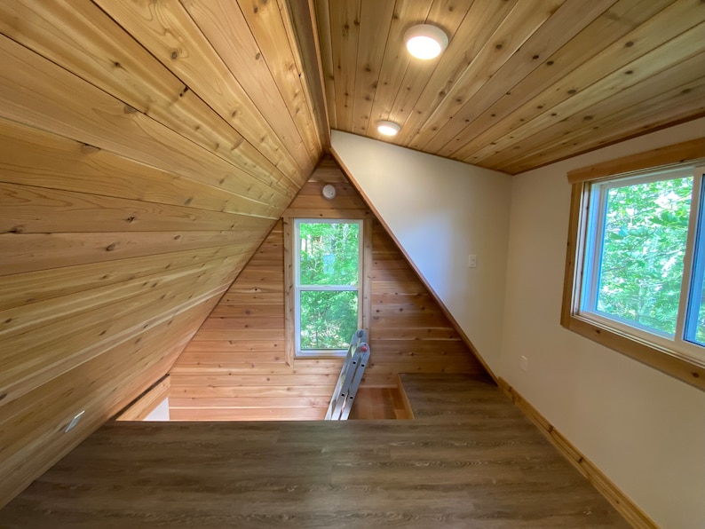 May include: A loft space with wood paneling on the walls and ceiling. There is a window with a view of trees outside. A ladder is leaning against the wall.